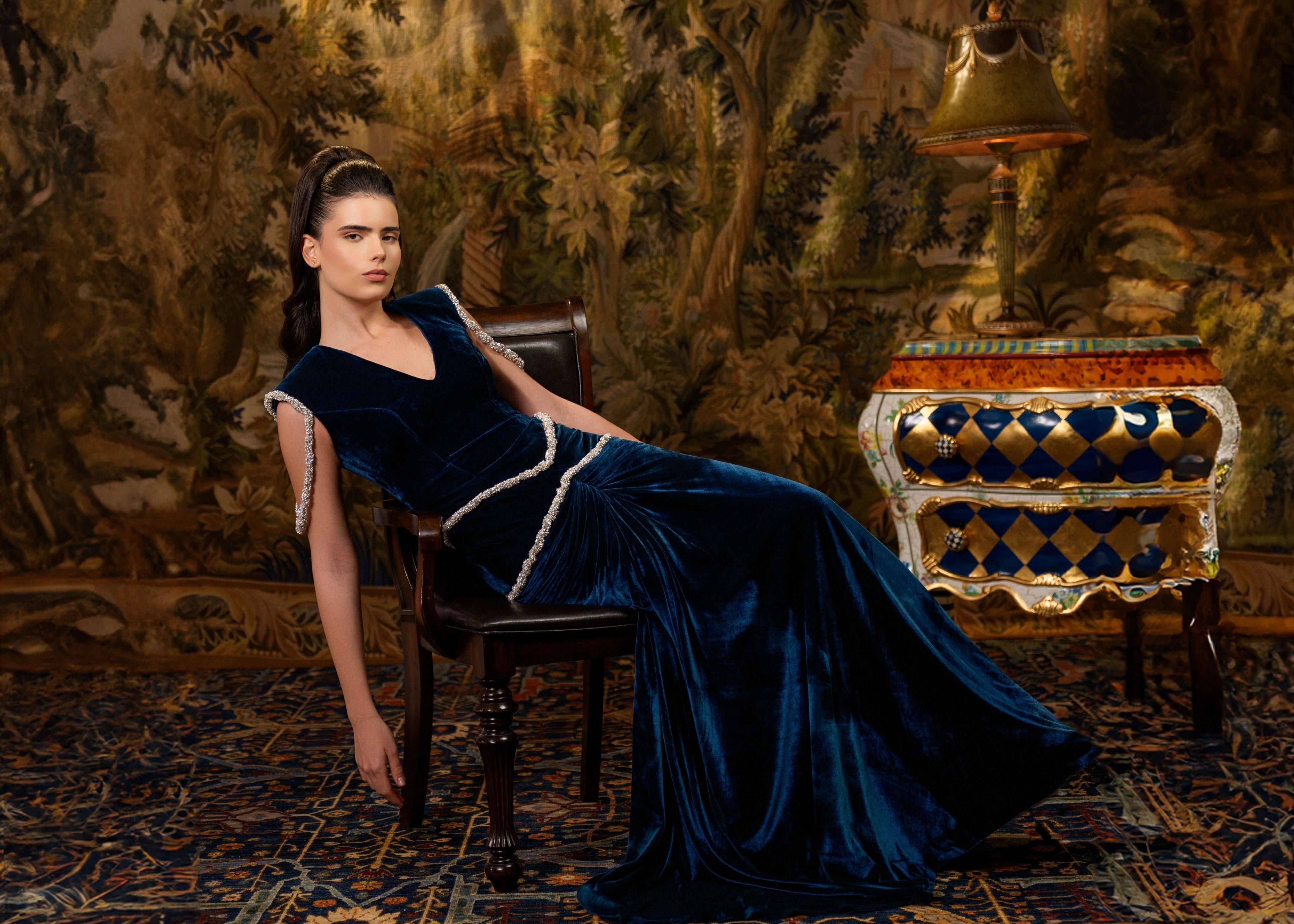 Woman in a blue evening gown sitting on a chair in an ornate room with intricate wall patterns and furniture.